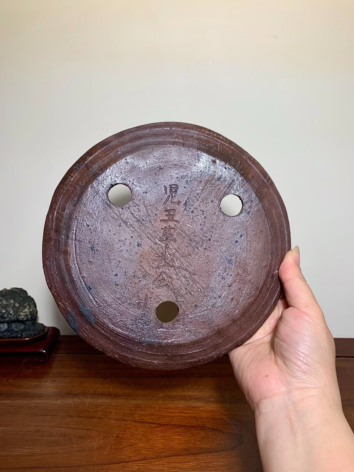 A hand holds a round, dark brown unglazed ceramic pot base with three holes and faded Japanese characters.