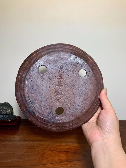 A hand holds a round, dark brown unglazed ceramic pot base with three holes and faded Japanese characters.