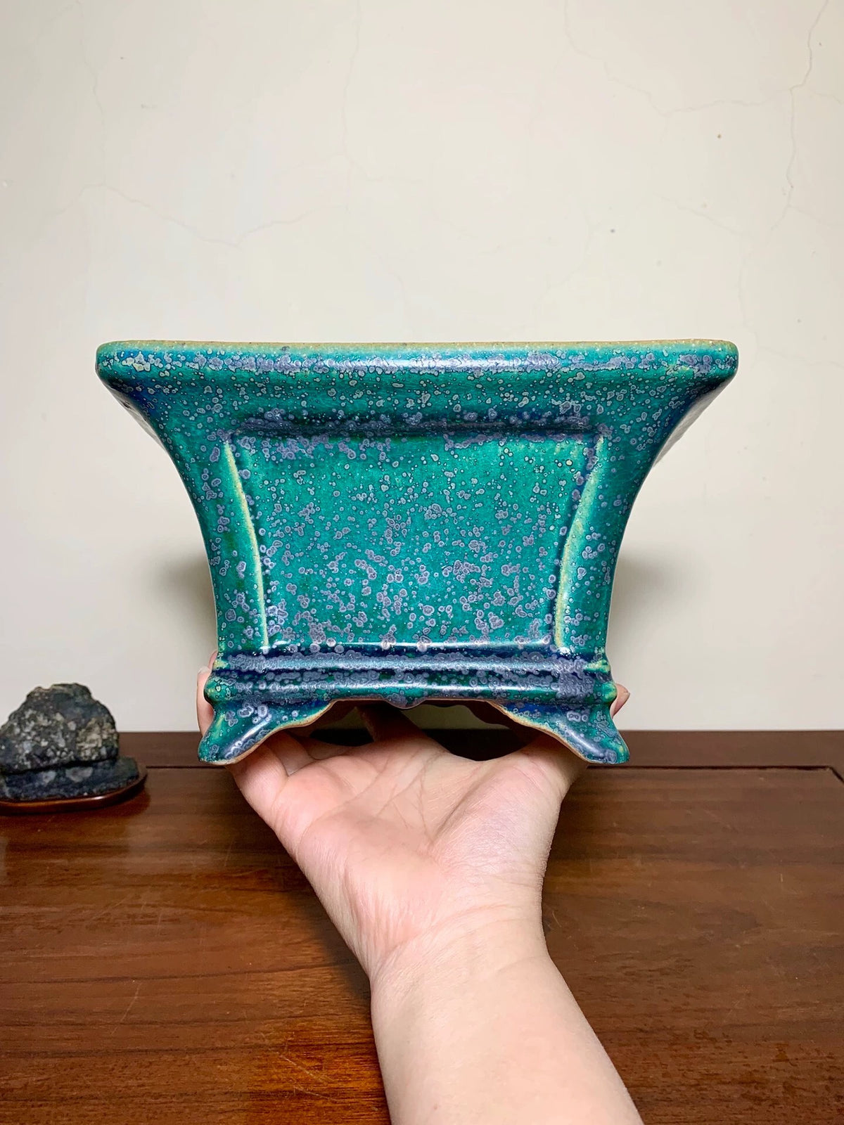 A hand holds a glossy, teal-glazed square pot with speckled blue and purple glaze.