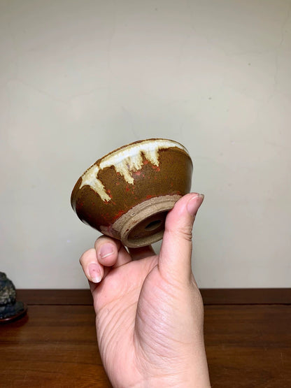 A hand holds a small, brown-glazed ceramic bowl with a white and red speckled rim.