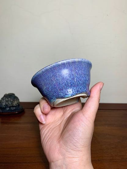 A hand holds a small, blue-glazed ceramic bowl with a speckled, glossy surface.