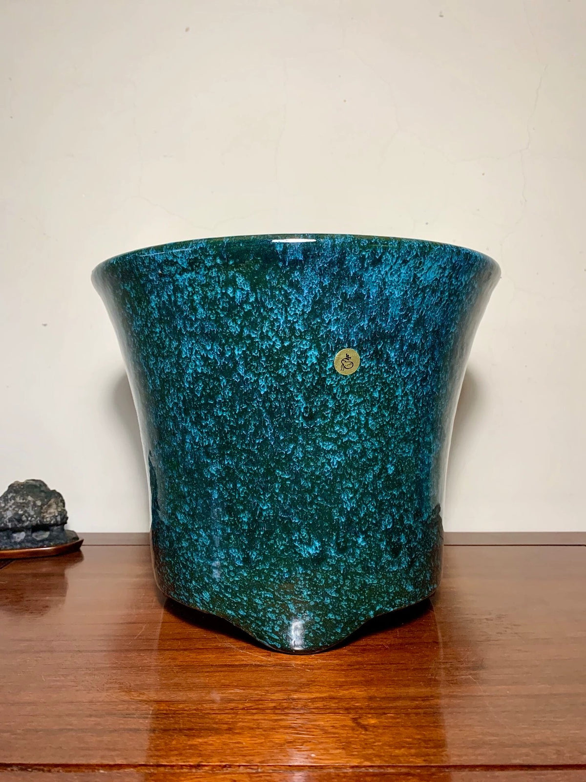 A glossy, speckled teal ceramic pot with a small gold mark, resting on a wooden surface.