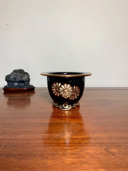 Black glazed pot with gold floral decoration and ornate foot, on wooden surface.