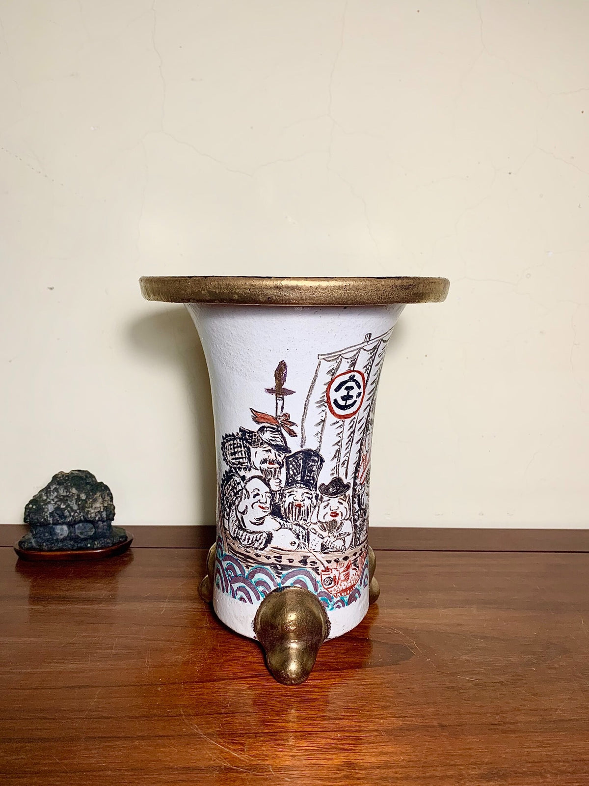 White ceramic pot with black and red painted figures, gold rim, and three brass feet.
