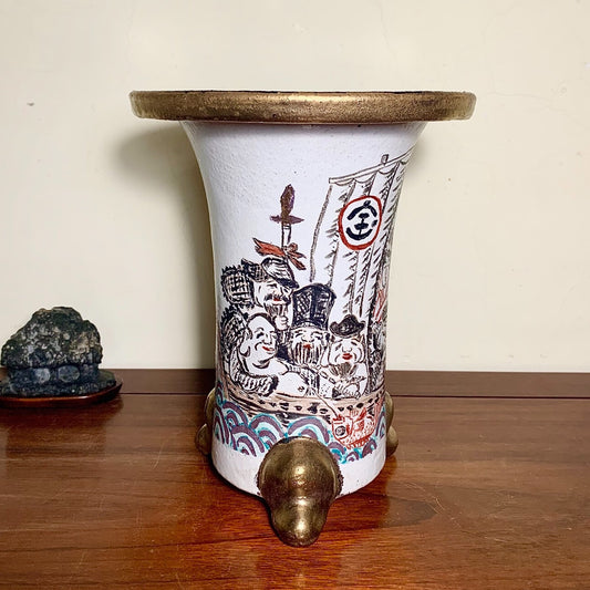White ceramic pot with black and red painted figures, gold rim, and three brass feet.