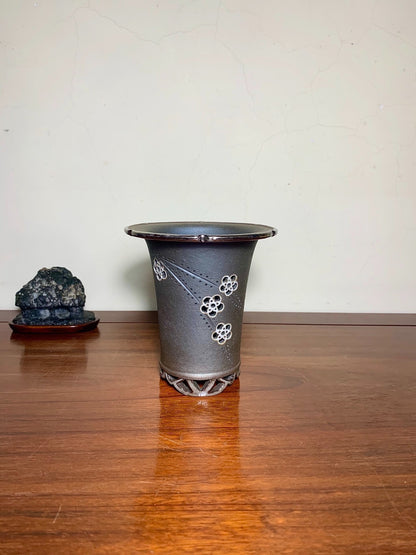 Dark grey glazed pot with white floral patterns and a perforated base, sitting on a wooden surface.