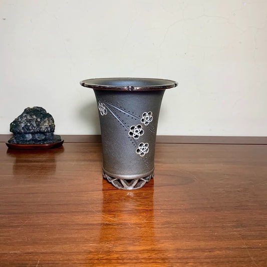 Dark grey glazed pot with white floral patterns and a perforated base, sitting on a wooden surface.