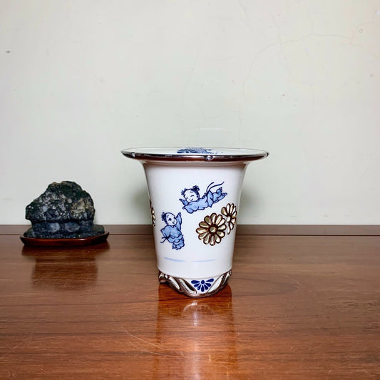 White ceramic pot with blue and brown painted figures and flowers, glossy finish.