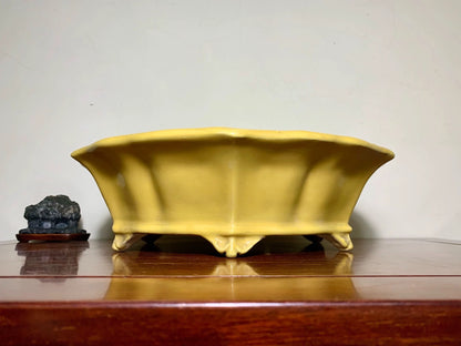 A matte yellow ceramic pot with flared sides and four feet, beside a dark stone on a wooden surface.