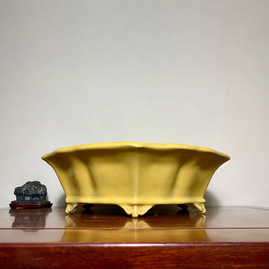 A matte yellow ceramic pot with flared sides and four feet, beside a dark stone on a wooden surface.