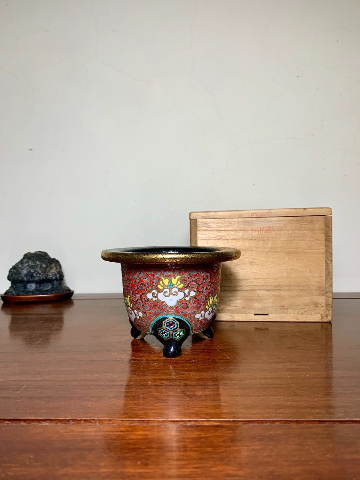A small, red-glazed pot with gold and white floral patterns, three black feet, sits on a wooden table.