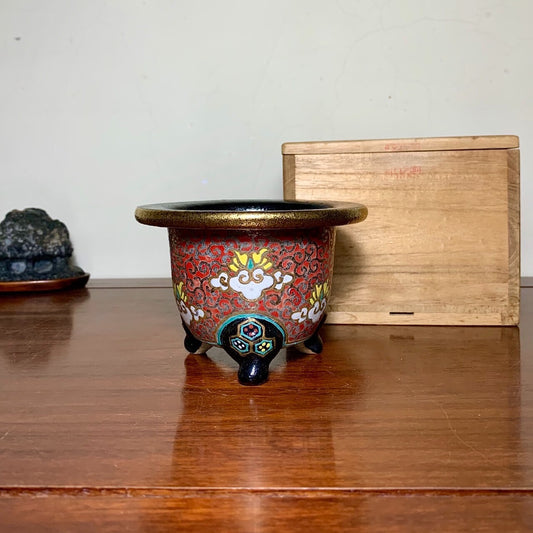 A small, red-glazed pot with gold and white floral patterns, three black feet, sits on a wooden table.