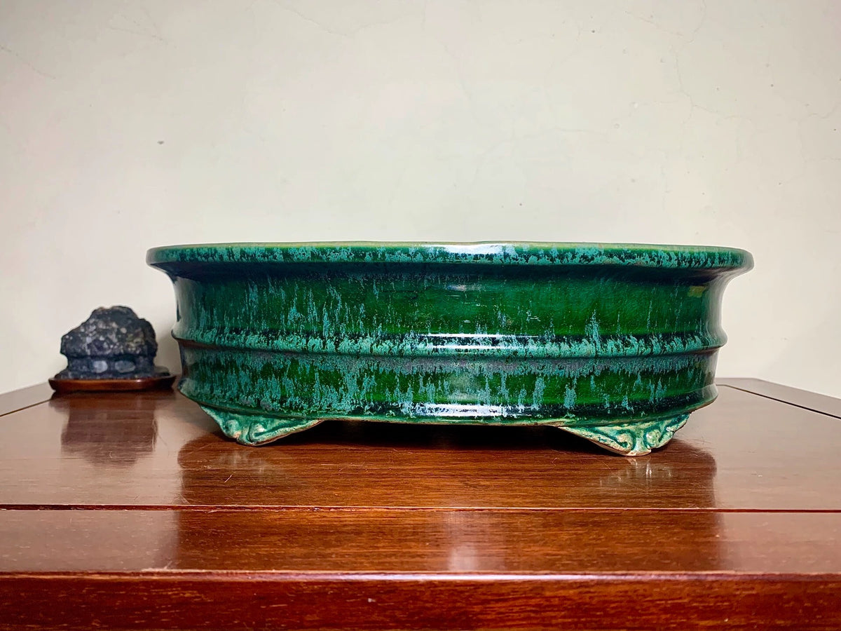 A glossy, deep green glazed ceramic pot with a wide rim and three feet, sits on a wooden surface.