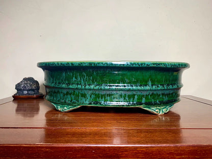 A glossy, deep green glazed ceramic pot with a wide rim and three feet, sits on a wooden surface.