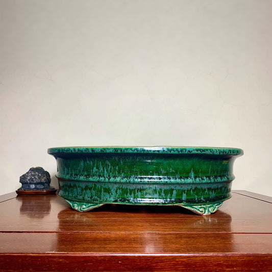 A glossy, deep green glazed ceramic pot with a wide rim and three feet, sits on a wooden surface.