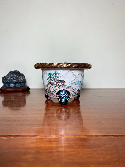 A small, light-colored ceramic pot with a painted landscape and a gold-trimmed rim.