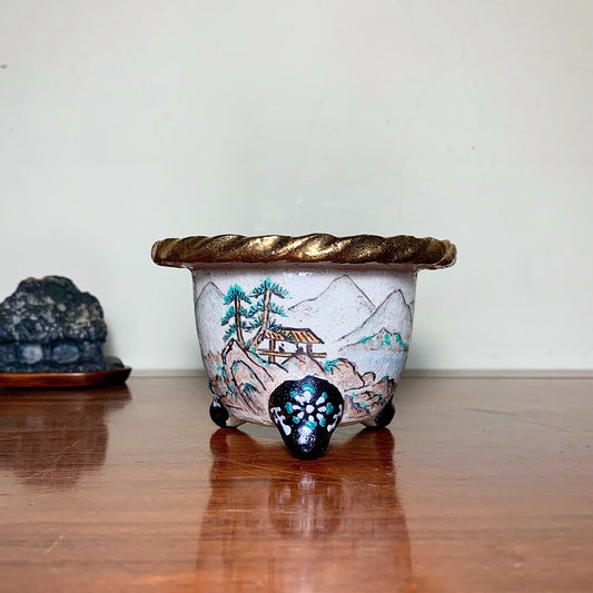 A small, light-colored ceramic pot with a painted landscape and a gold-trimmed rim.