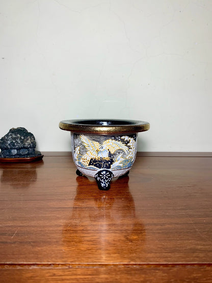 A glazed ceramic pot with a black rim, gold and white phoenix design, and three ornate feet.