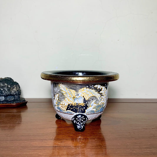 A glazed ceramic pot with a black rim, gold and white phoenix design, and three ornate feet.