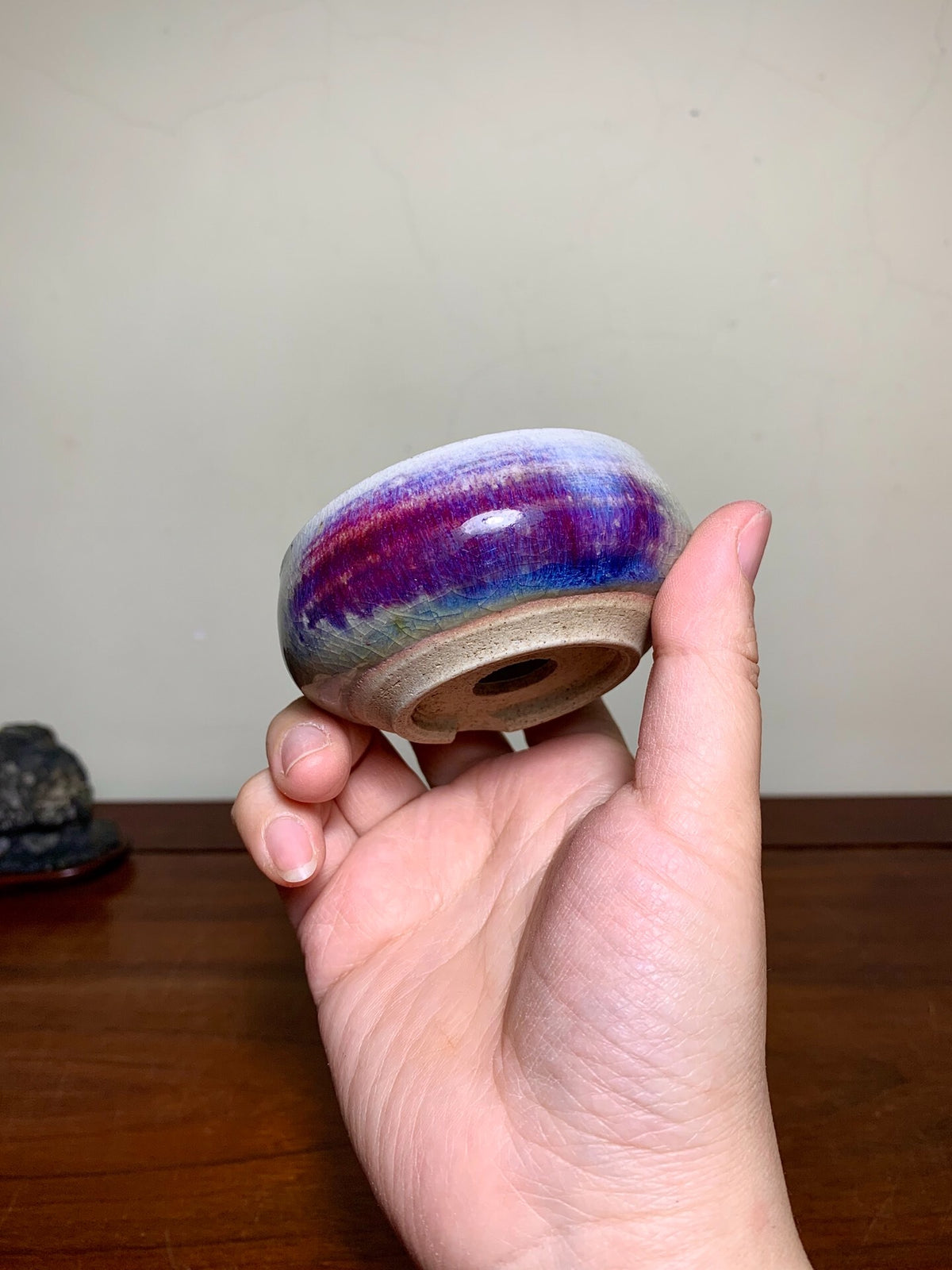 A hand holds a small, glazed ceramic pot with a vibrant purple and blue rim.