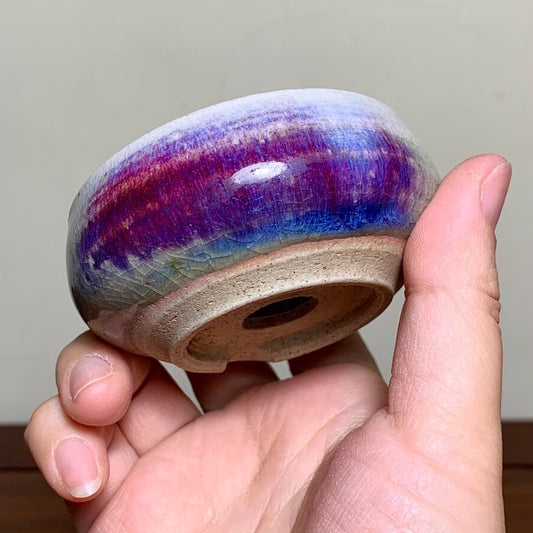A hand holds a small, glazed ceramic pot with a vibrant purple and blue rim.