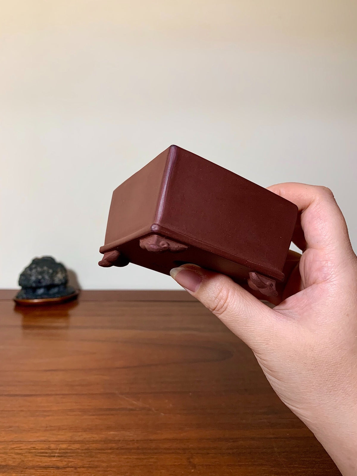 A hand holds a small, matte brown square pot with four small feet, unglazed ceramic.