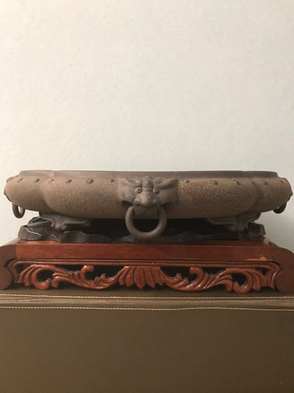 Dark brown glazed pot with lion-head ring handle, resting on carved wooden stand.