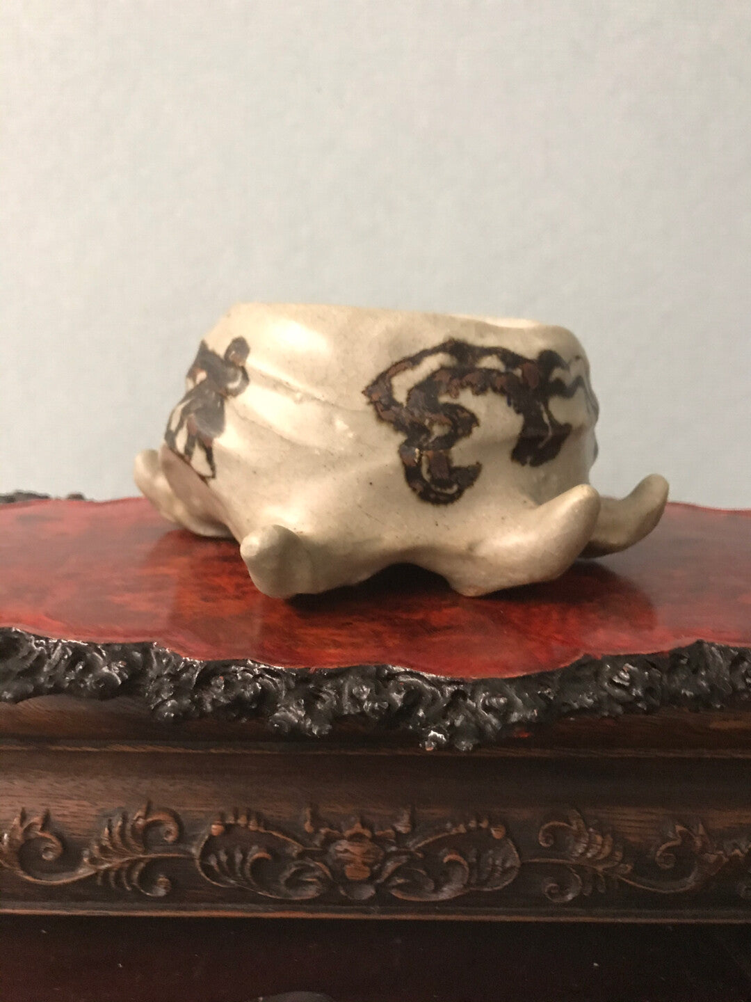 A small, off-white ceramic pot with dark brown abstract markings rests on a carved wooden stand.