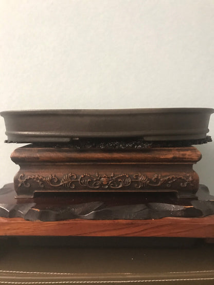 Dark brown glazed ceramic pot on ornate wooden stand with carved floral pattern.