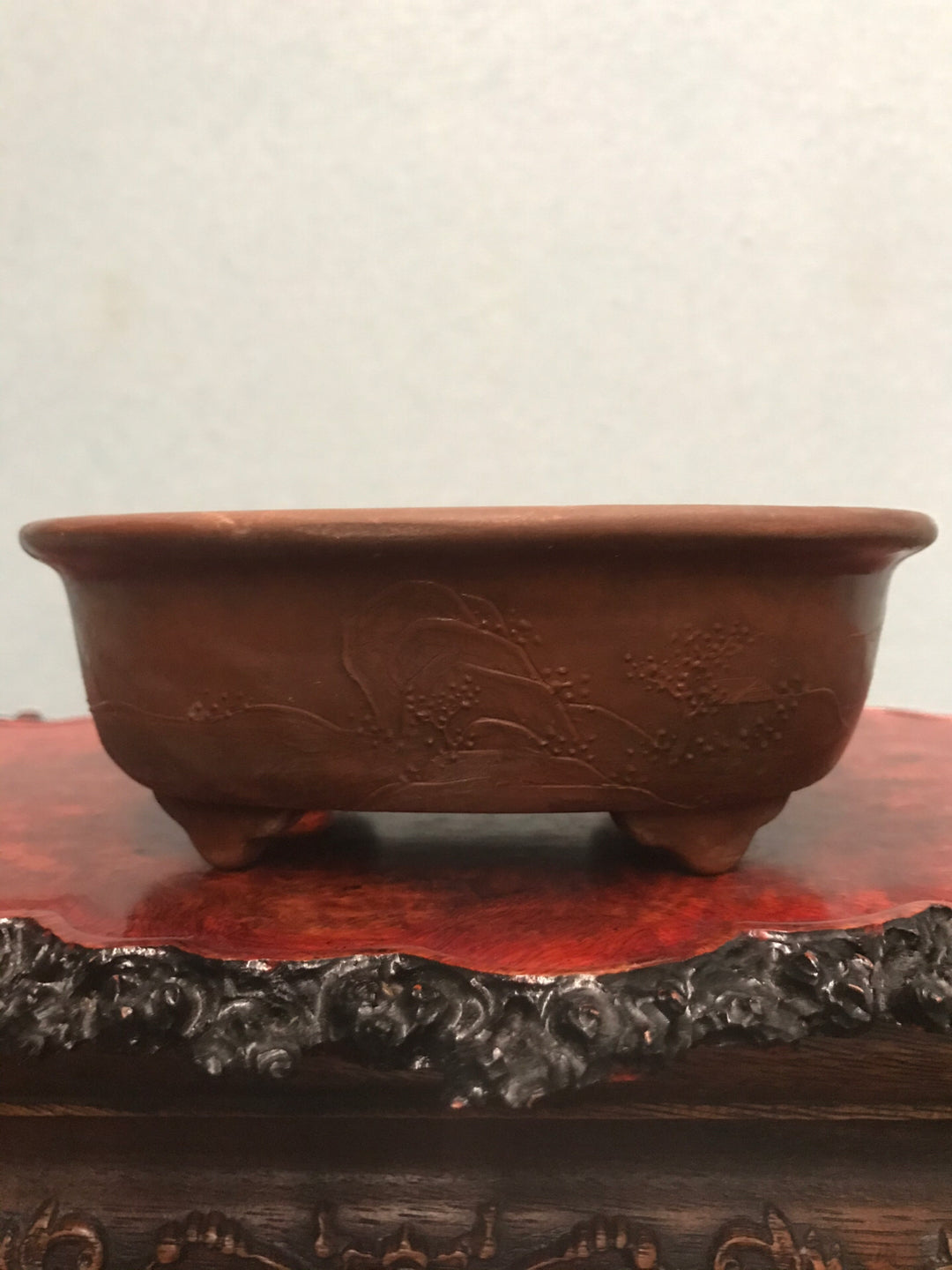 A brown, unglazed ceramic pot with carved floral patterns rests on a red lacquered stand.