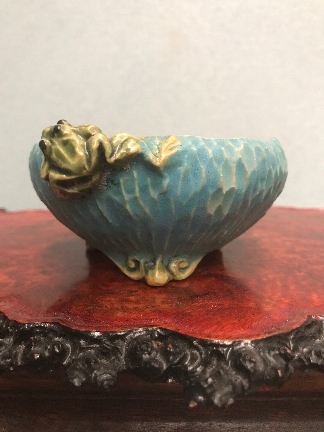 A glazed blue ceramic bowl with a green frog figurine on its rim, resting on a red wooden stand.