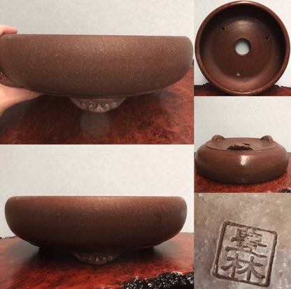 A brown, unglazed ceramic bonsai pot with a wide rim, three feet, and a square maker's mark.