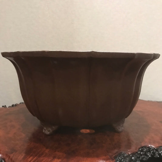A dark brown, fluted ceramic pot with three feet and drainage holes, showing an engraved mark on its base.