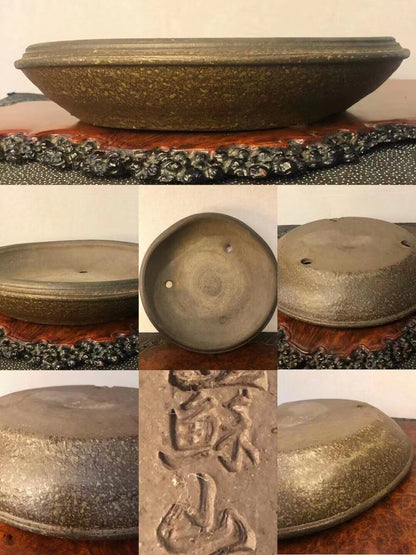 A dark, speckled glazed pot with three drainage holes and carved characters on its base.