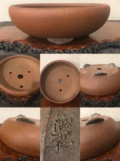 A round, brown, unglazed ceramic pot with three drainage holes and an incised signature.