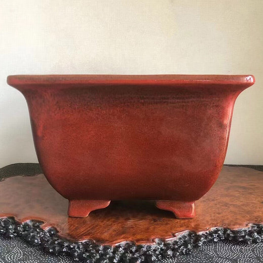 A square red glazed pot with a glossy finish and a small central hole in the base.
