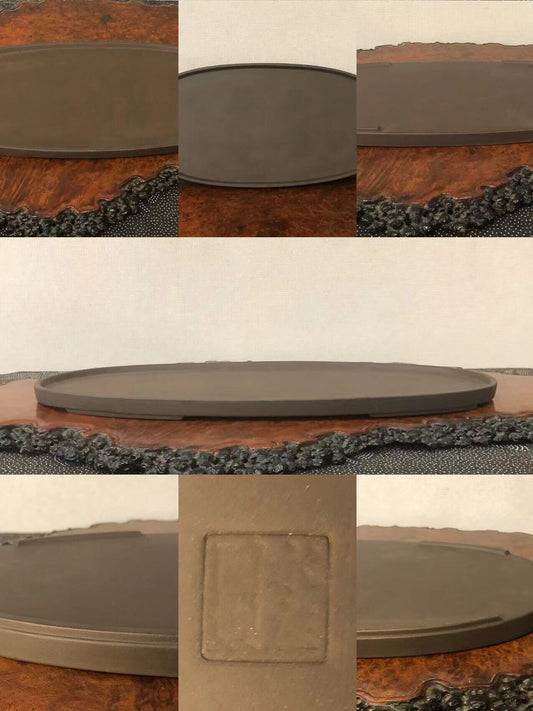 A dark grey, oval-shaped, unglazed ceramic bonsai pot with a square maker’s mark.