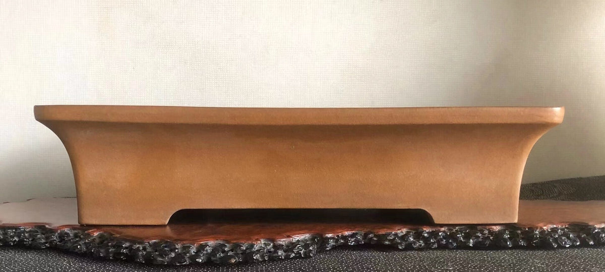 A matte, light brown, rectangular bonsai pot with curved ends and a central foot rests on a dark, textured base.
