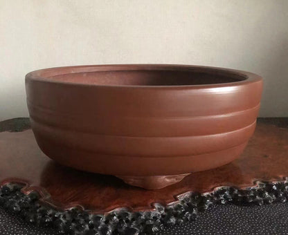 A wide, reddish-brown ceramic pot with horizontal ridges and three feet, resting on a dark wooden stand.