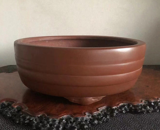A wide, reddish-brown ceramic pot with horizontal ridges and three feet, resting on a dark wooden stand.