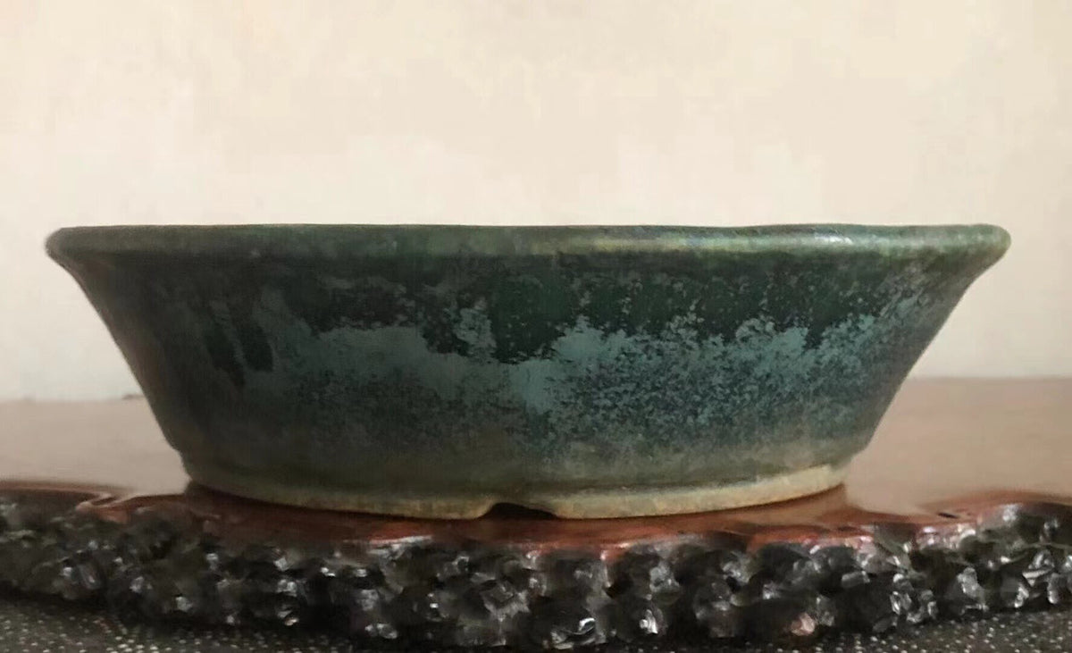 A wide, shallow glazed pot with a mottled green and blue glaze sits on a wooden stand.