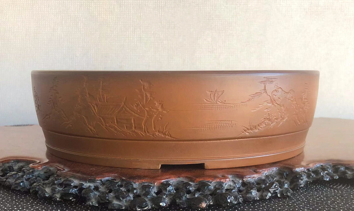 A brown glazed pot with carved landscape scenes on its rim.
