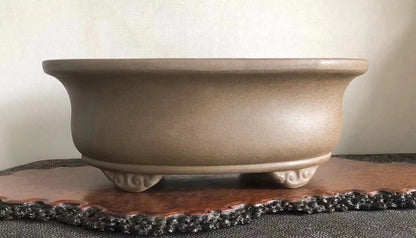 A beige, matte-finished ceramic pot with three ornate feet, resting on a wooden tray.