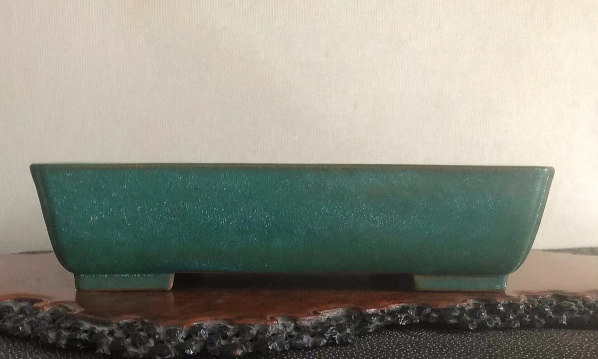 A matte teal ceramic pot with a rectangular shape and slightly curved sides rests on a wooden stand.