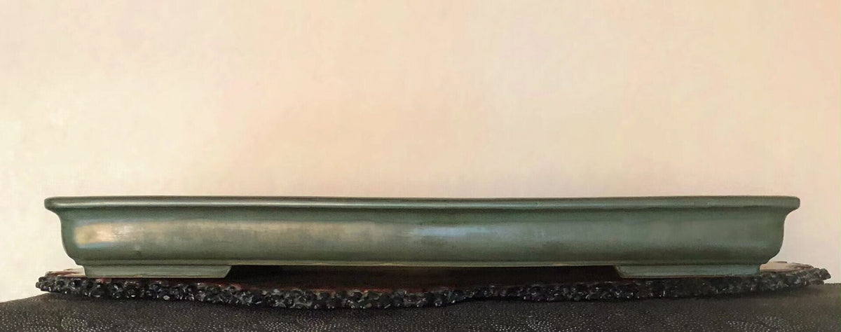 A long, low, olive-green glazed pot rests on a dark, textured stand.