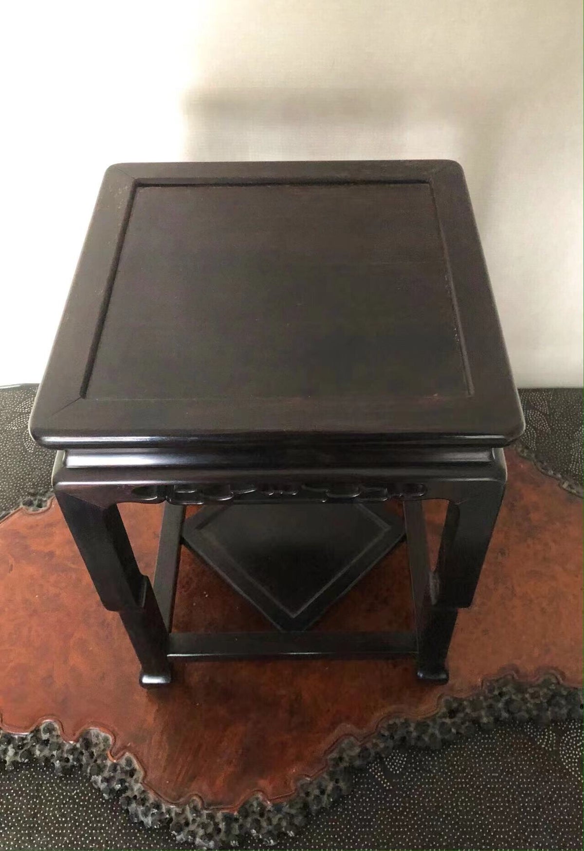 Dark wooden stand with square top and four legs, glossy finish, sits on patterned rug.