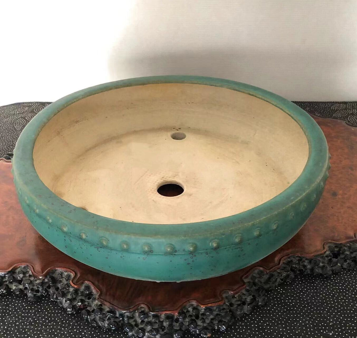 A round, teal-glazed pot with a dotted rim and two holes, resting on a wooden stand.