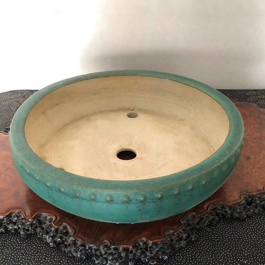 A round, teal-glazed pot with a dotted rim and two holes, resting on a wooden stand.