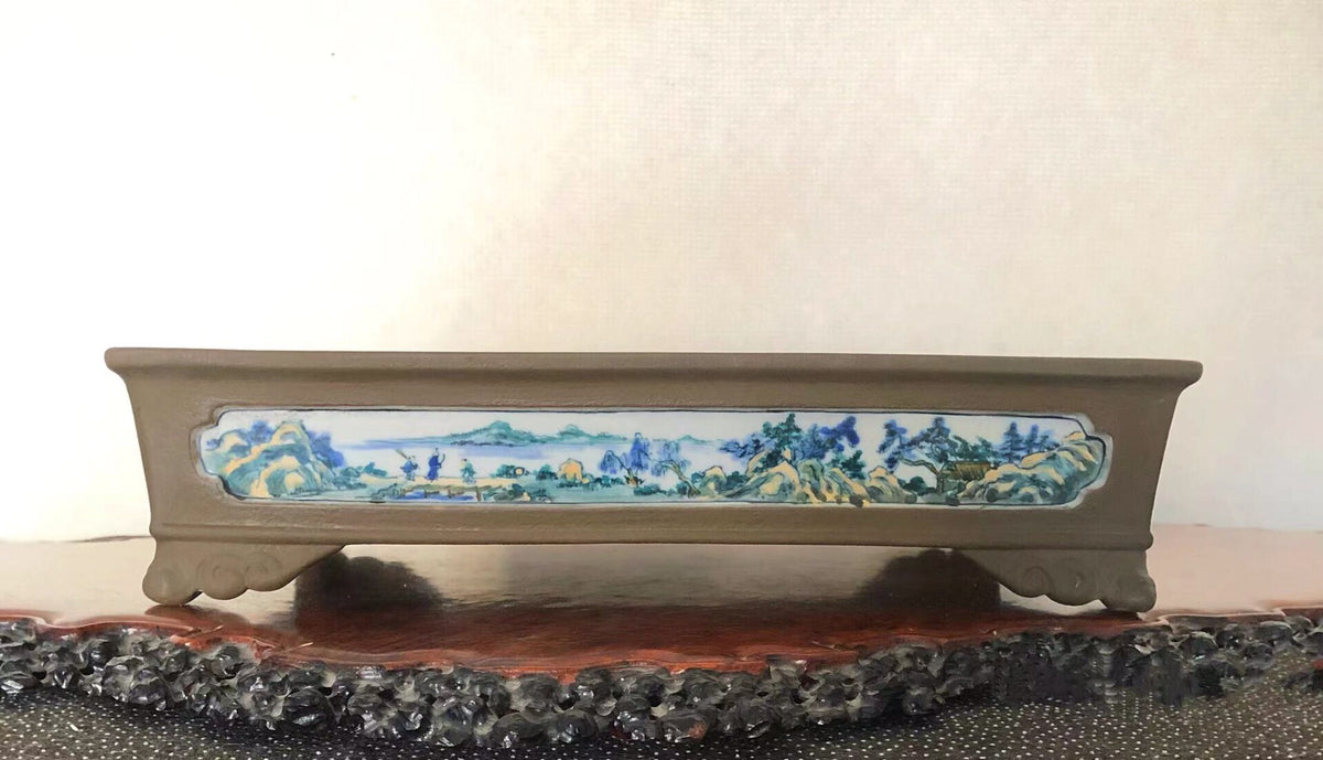 A long, brown glazed pot with a painted landscape scene on its front panel.