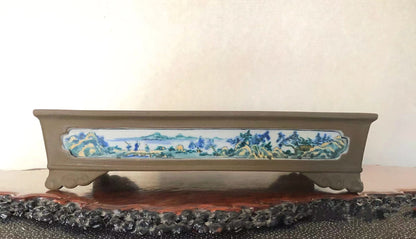 A long, brown glazed pot with a painted landscape scene on its front panel.